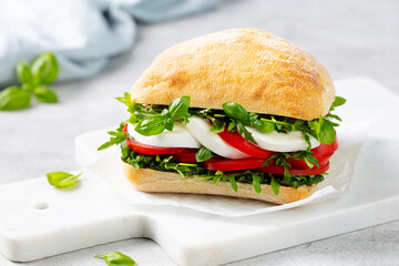 Ciabatta sandwich with mozzarella, tomatoes, pesto sauce and fresh basil on marble serving board. Light grey background. Close up.