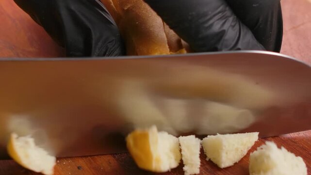 A sharp knife cuts a fresh bread roll on a wooden board. The bread is diced into small, even cubes to be baked into crispy, golden homemade croutons for salads or soups