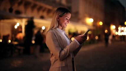 Smiling woman in beige suit holding smartphone at night, reading message with joy while standing on illuminated city street - Powered by Adobe