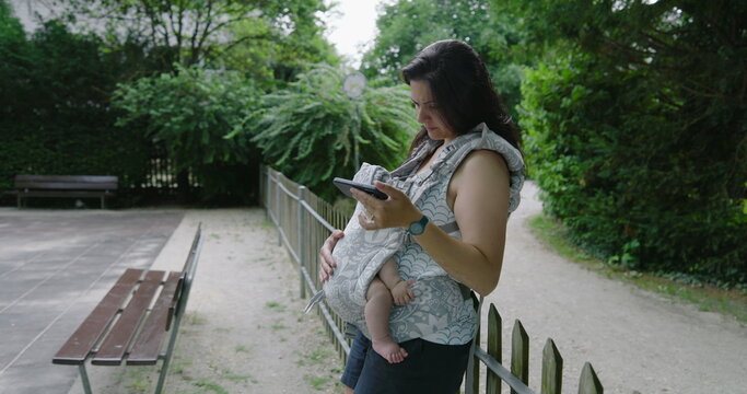Mother with a preoccupied expression, holding her baby in a front carrier while intently focusing on her smartphone in a park, highlighting the blend of modern concerns and parenting - Powered by Adobe