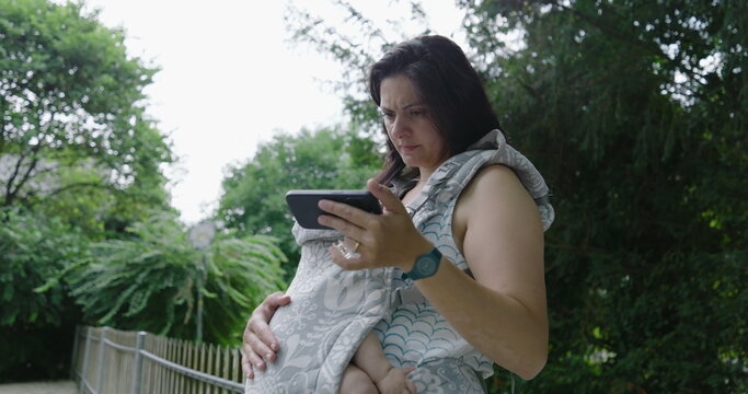 Mother with a preoccupied expression, holding her baby in a front carrier while intently focusing on her smartphone in a park, highlighting the blend of modern concerns and parenting