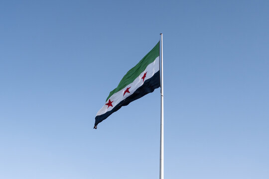 The official Syrian flag adopted in December 2024, with green, white, and black stripes and three red stars, waving against a clear blue sky in the Syrian capital, Damascus.