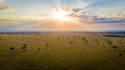 Golden sun rays stream through dramatic clouds over a vast savanna landscape dotted with acacia trees at sunset