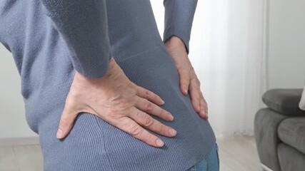 Back pain. Middle aged woman holding her lower back and massaging it with her hands at home. Close-up, rear view, side view. Lumbago, sciatica, lower back pain, lumbago in the back, hernia, protrusion - Powered by Adobe