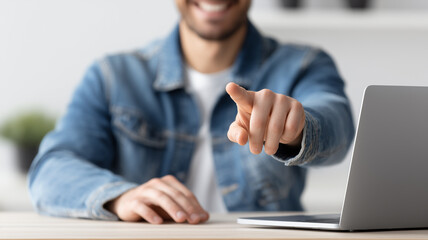 Smiling person pointing toward the viewer near a laptop symbolizing call to action choice and the concept of user engagement and recruitment.
