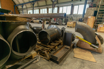 Large industrial metal pipe fittings and components stored in a workshop, showing steel elbows and cylinders on the floor. Concept of heavy industry, metalworking, and manufacturing equipment