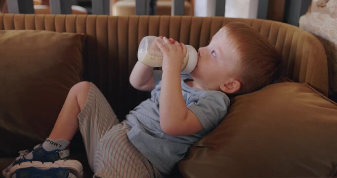 A little boy drinking milk from child bottle lying on sofa
