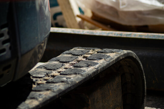 A detailed close-up of a dirty rubber track from a mini excavator. The worn tread is caked with mud, highlighting its rugged nature and use on rough terrain at an active urban job site.
