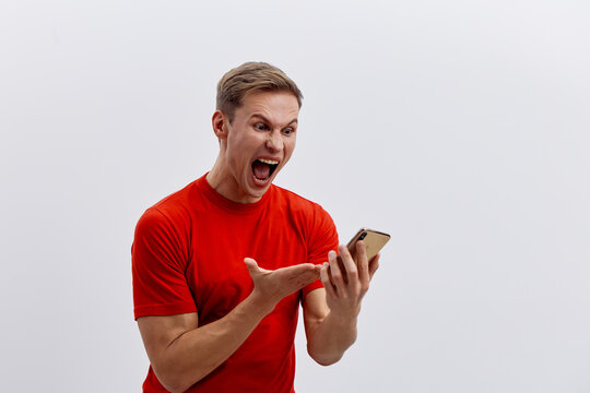 Excited young man with short blond hair in a bright red t-shirt shouting happily while looking at his smartphone with a joyful expression isolated on a plain white background. People lifestyle concept