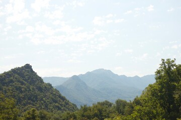 the mountain landscape with green hills covered with forest on the forefront and the bluish mountains on the background under the light blue sky on a sunny day