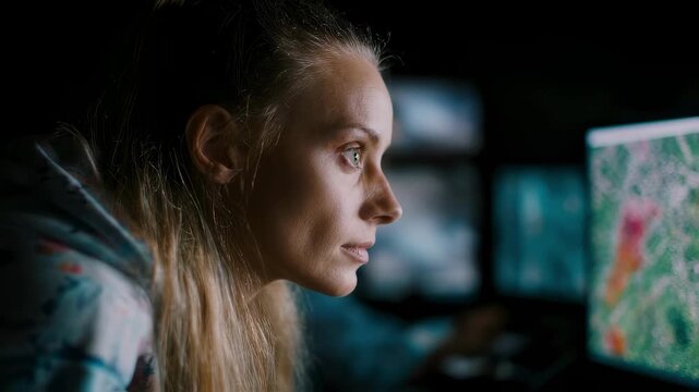 Concentrated volunteer in medium shot reviewing satellite imagery on monitor face and screen in sharp focus darkened workspace and map tiles out of focus to highlight task.