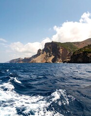 Rugged coastline meets a tranquil ocean.  Dark blue water lapping at a light tan/brown cliff face.  Clouds drift above