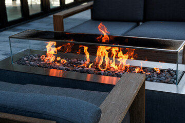 modern outdoor fire pit with glowing flames over stones, surrounded by seating for relaxation and gathering, cozy fireplace setting © Veronique