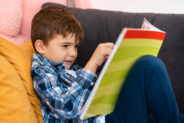 A happy child boy sitting on a sofa at home and reading a book.