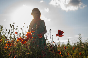Woman in a green dress gently holding poppies in a sunlit field, silhouetted against a bright sky. Captures summer beauty, nature, and tranquility