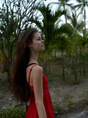 young woman with long hair wearing red dress standing outdoors in tropical garden, side profile portrait with natural greenery and soft lighting, peaceful expression
