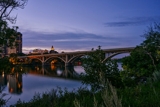 Golden sunset view of Saskatoon with a bridge crossing the river and high-rise buildings in the background.