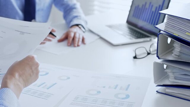 Office workers or auditors are analyzing financial reports and checking documents while sitting at white table with a stack of blue folders in the foreground. Audit and taxes theme in business - Powered by Adobe