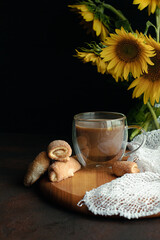 Coffee in a cup on a wooden board with fresh sugar cookies. On a dark background bright yellow sunflowers in a vase.