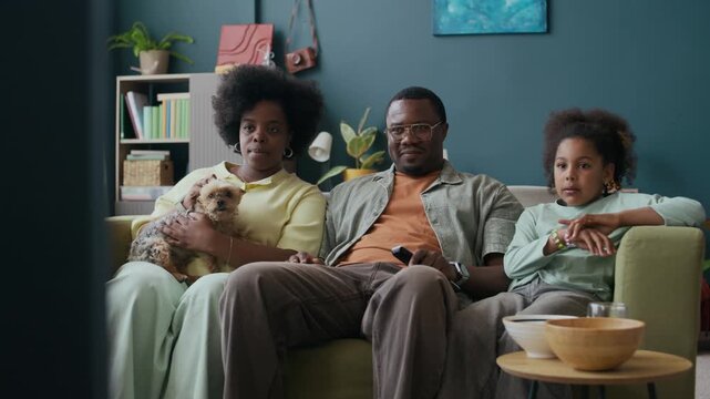 Family of three sitting on couch in living room, pointing at screen of TV set and discussing broadcast of sports game or documentary moments while spending evening together