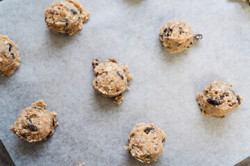 Close up of round raw oatmeal and müsli raisin cookie dough scoops on cookie sheet with baking paper made of flour, rolled oats, sugar, eggs, raisins, muesli, baking soda and powder before baking