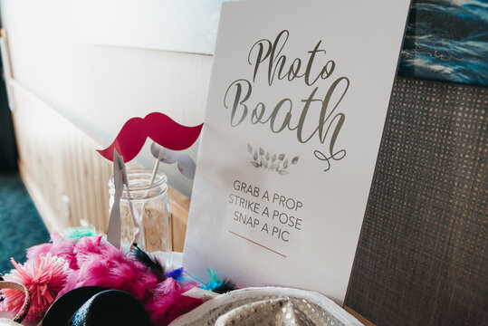 wedding photo booth setup with props in a clear mason jar, sunlight streaming through a window, decorative sign reading &ldquo;Photo Booth. Grab a prop, strike a pose, snap a pic,&rdquo; festive party scene
