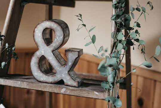 Rustic wedding d&eacute;cor with metal ampersand symbol on a ladder surrounded by greenery, romantic decorative detail for ceremony or reception, symbolic love and unity decoration