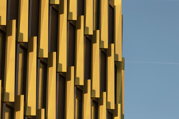 A closeup image highlights the innovative design of a contemporary building facade with vertical, protruding elements against a clear blue sky, enhancing the urban landscape