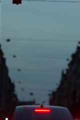 The rear light of a parked car shines brightly as it patiently waits at a traffic signal, nestled among busy city streets, during the evening hours, all beneath an overcast sky