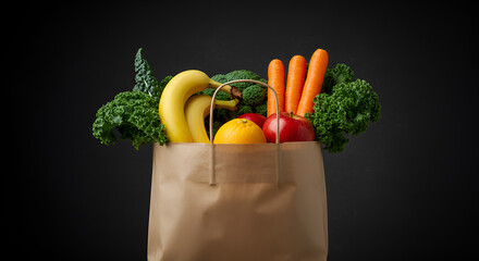 Paper bag filled with fresh vegetables and fruits such as broccoli, lettuce, oranges, apple, cucumber, and strawberries on black background.