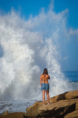Fototapeta premium PORTRAIT: Young woman is admiring a huge wave crashing against rugged rocky Moroccan shore sending up a large white splash of foam and spray. Beach walk with dramatic scenery of mighty Atlantic Ocean