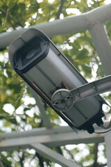 Close-up of an outdoor security camera mounted on a metal pole. CCTV surveillance equipment with a blurry green tree background.