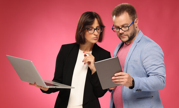 businessman and businesswoman looking at tablet consulting deciding. man and woman in business clothes with tablet and laptop on pink background
