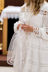 Close-up of a woman in a white lace dress holding a decorated christening candle, rosary and veil,...