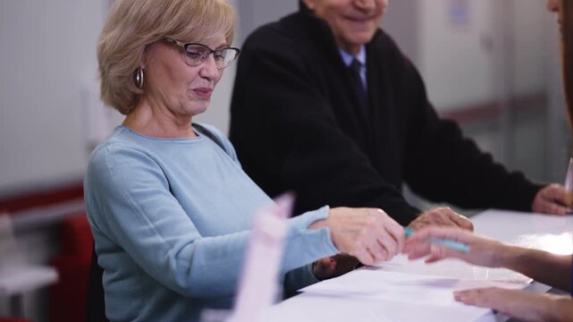 A senior woman signs medical forms at a reception desk in a clinic.