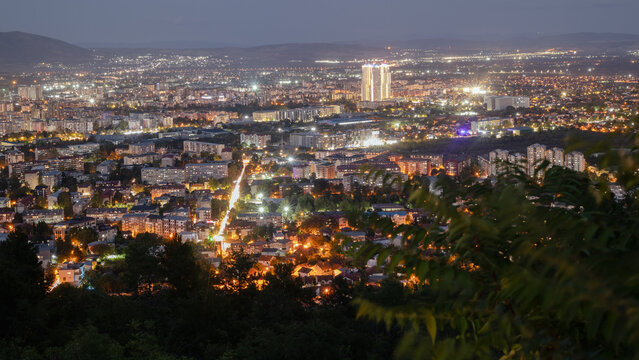 an aerial view of the city of skopje at night, showing the urban landscape with illuminated buildings and streets under a twilight sky. the city lights create a vibrant and warm glow