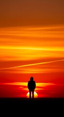 Fototapeta premium A silhouetted figure stands alone, gazing at a vibrant, fiery sunset. The intense orange and red hues dominate the sky, creating a dramatic and contemplative scene.
