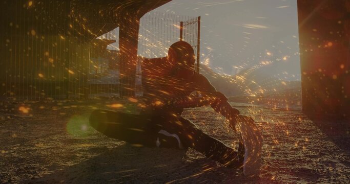 Dancing man in hoodie weaving spark-like lights on gravel under overpass pillars, fence at dusk - Powered by Adobe