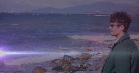 Gazing man in green coat and sunglasses on rocky shore, cargo ships, lens flare, copy space