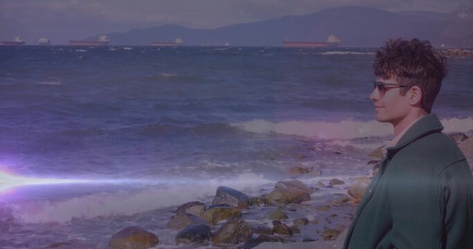 Gazing man in green coat and sunglasses on rocky shore, cargo ships, lens flare, copy space - Powered by Adobe