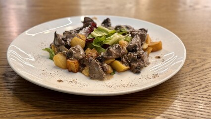 Warm salad with chicken liver in cream sauce with fried potatoes, mushrooms and mixed green salad in a white porcelain plate on a brown wooden table in an elite restaurant