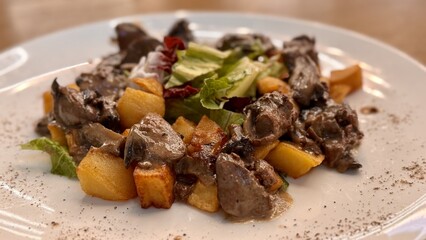 Warm salad with chicken liver in cream sauce with fried potatoes, mushrooms and mixed green salad in a white porcelain plate on a brown wooden table in an elite restaurant