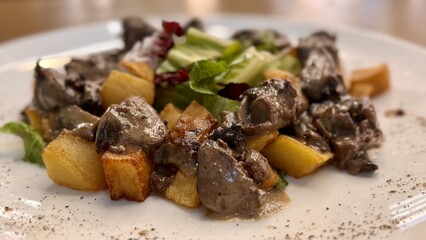 Warm salad with chicken liver in cream sauce with fried potatoes, mushrooms and mixed green salad in a white porcelain plate on a brown wooden table in an elite restaurant