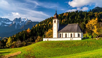 Fototapeta premium A picturesque church stands amidst autumnal foliage, nestled against a backdrop of snow-capped mountains under a vibrant sky.