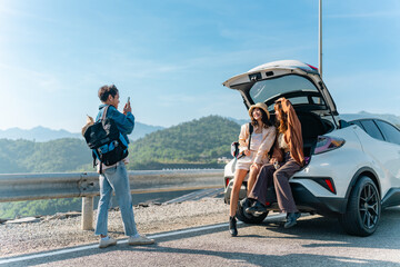 Happy Young Asian man and woman road trip travel by EV car countryside on summer holiday vacation. Generation z people friends sitting on car trunk and using mobile phone taking picture together. © CandyRetriever 