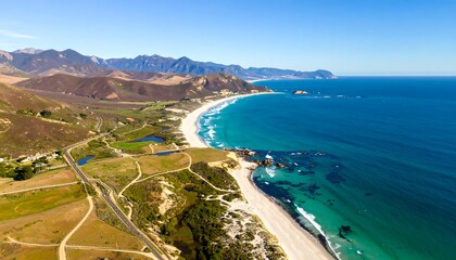 Coastal aerial with beach, clear water, and hills under a blue sky