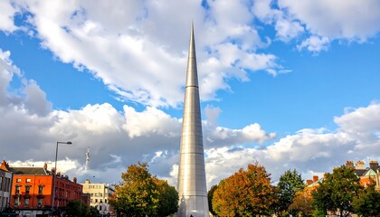 A tall, modern, pointed monument stands prominently against a vibrant sky filled with fluffy clouds, surrounded by autumn-hued trees and urban buildings.