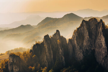 Majestic mountain range during sunrise, golden light on rocky cliffs, mist in valleys, dramatic natural background.
