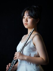Teen girl playing the flute in a white dress with soft lighting and a dark background. © Yuanxing
