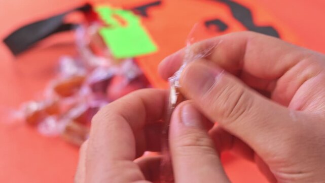 Hand taking and opening wrapped candies from orange felt jack-o'-lantern bag with an orange background. 